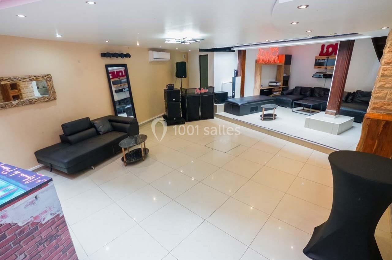 Salle lumineuse avec canapés noirs, tables basses, miroir mural et espace ouvert sur un sol en carrelage clair.