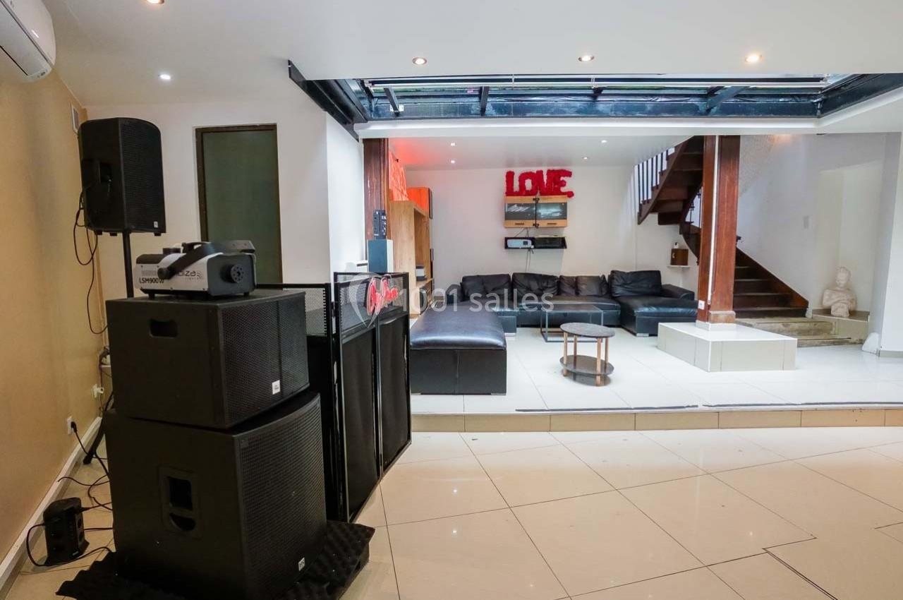 Salon moderne avec canapé en cuir noir, table basse, système audio et éclairage naturel provenant d'une verrière.