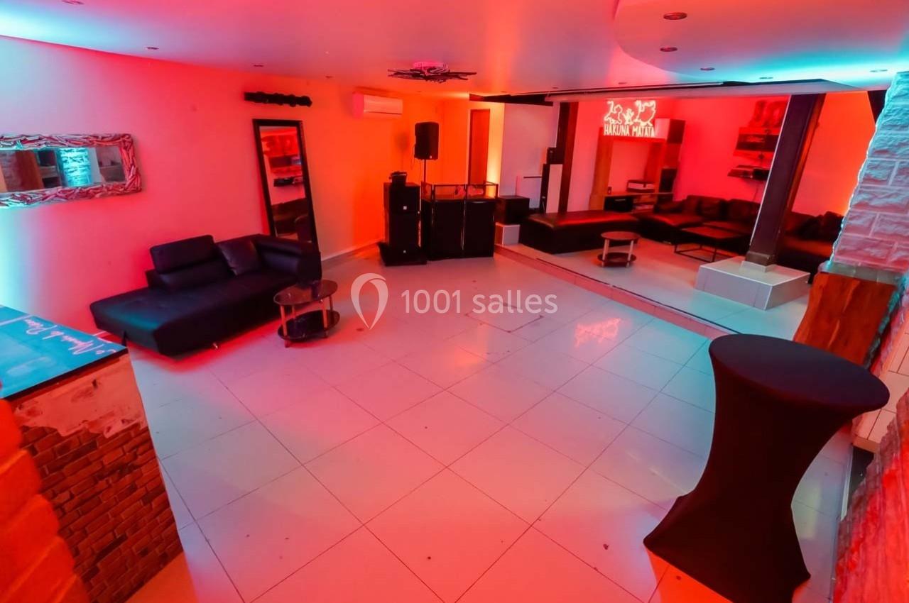 Salle moderne avec éclairage coloré, canapés noirs, tables basses et espace dégagé sur un sol carrelé blanc.