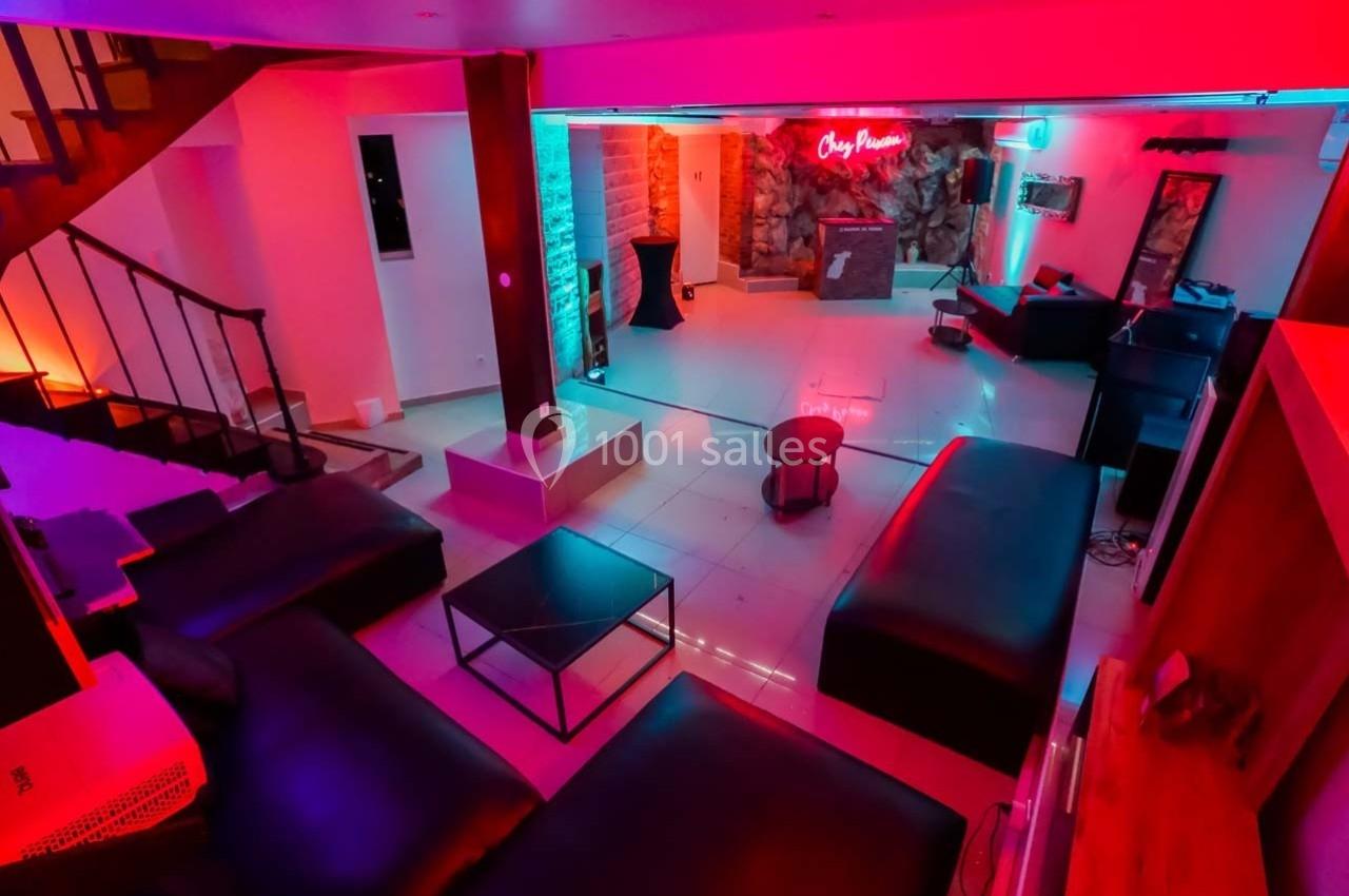 Salle moderne avec éclairage néon coloré, canapés noirs, tables basses et escalier en bois visible à gauche.