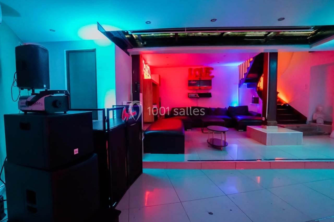 Salle moderne éclairée par des lumières colorées, avec canapés, table basse, escalier et équipement audio.