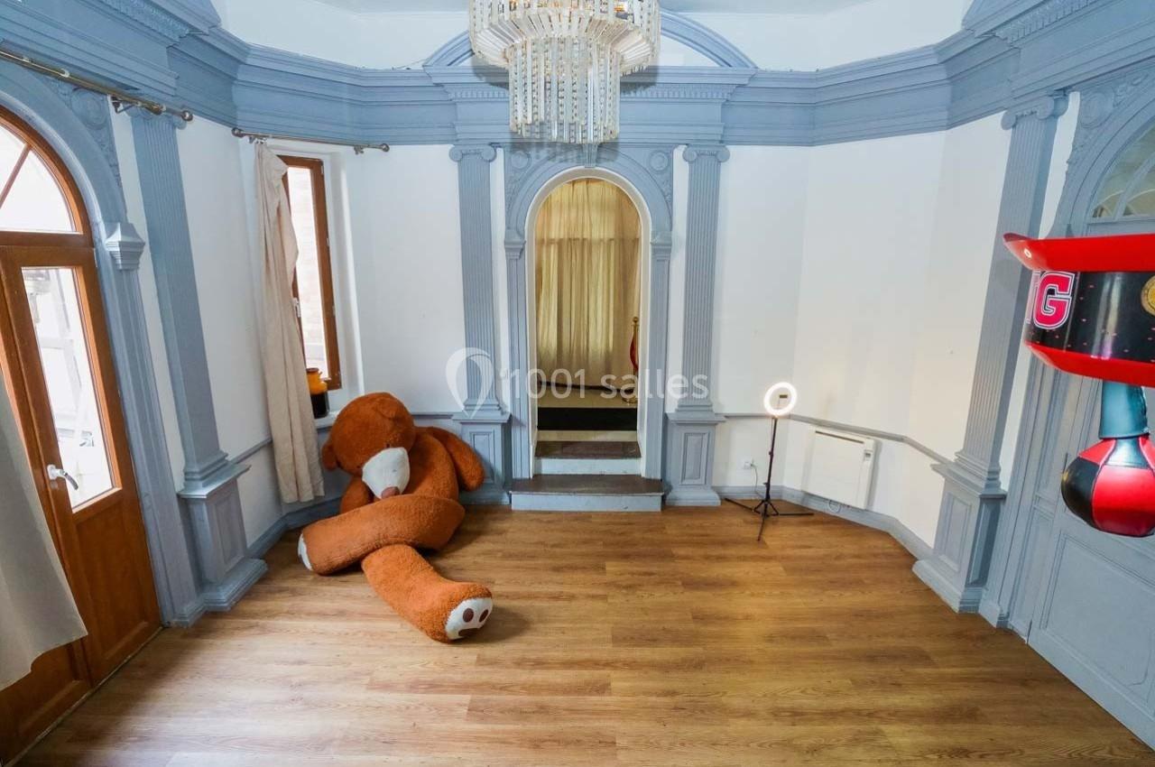 Une pièce lumineuse avec parquet, murs blancs et moulures grises, un grand ours en peluche posé au sol.