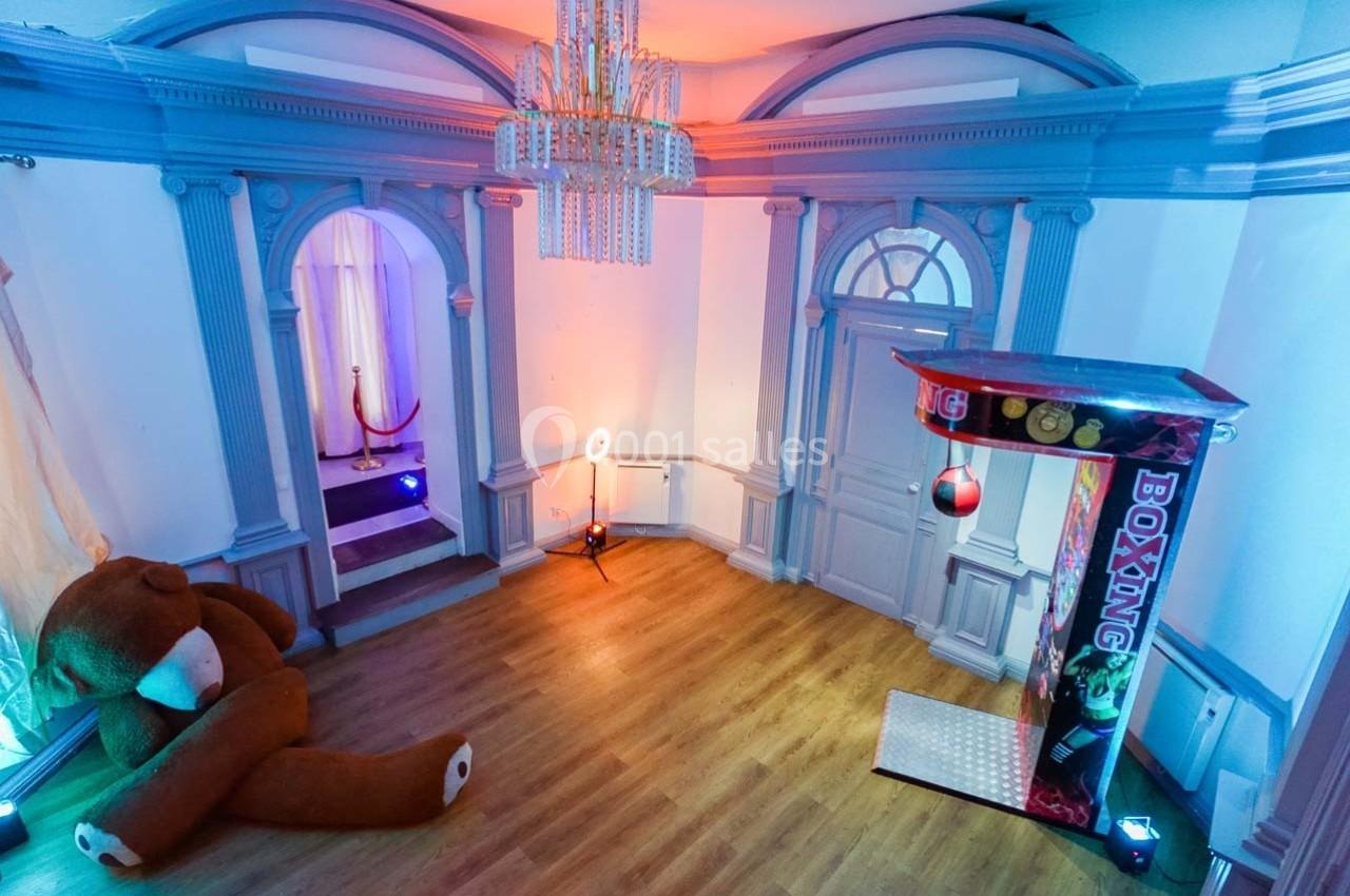Salle lumineuse avec un sol en bois, un grand ours en peluche, un jeu de boxe et des arches décoratives.