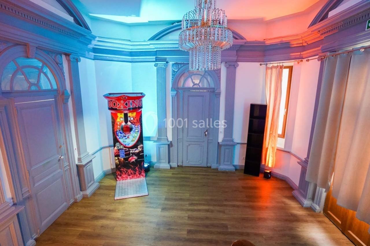 Salle lumineuse avec parquet, murs ornés de moulures, un lustre et une machine de jeu de boxe près d'une fenêtre.