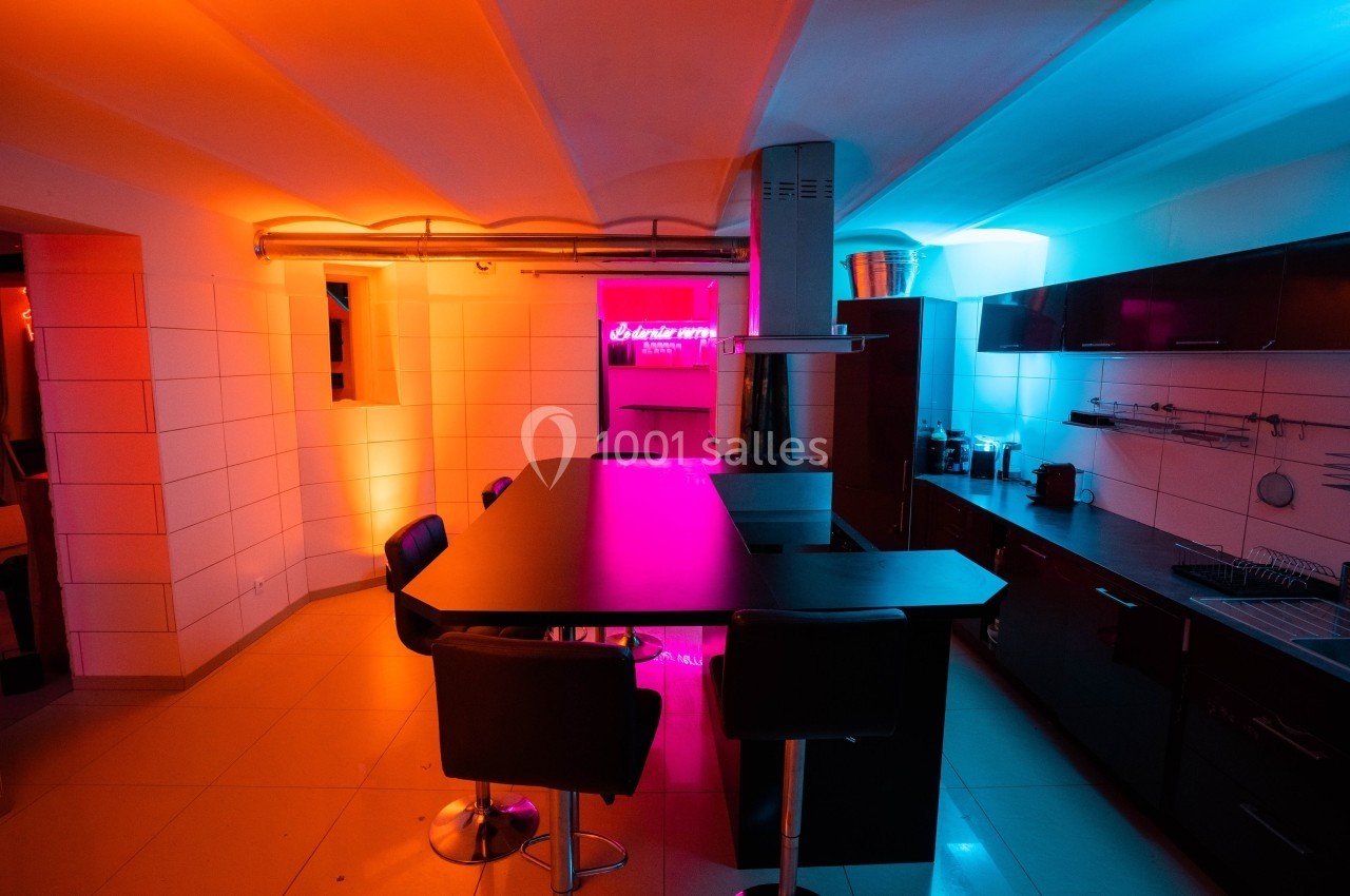 Cuisine moderne avec éclairage néon coloré, comptoir central noir et chaises hautes dans un espace carrelé.