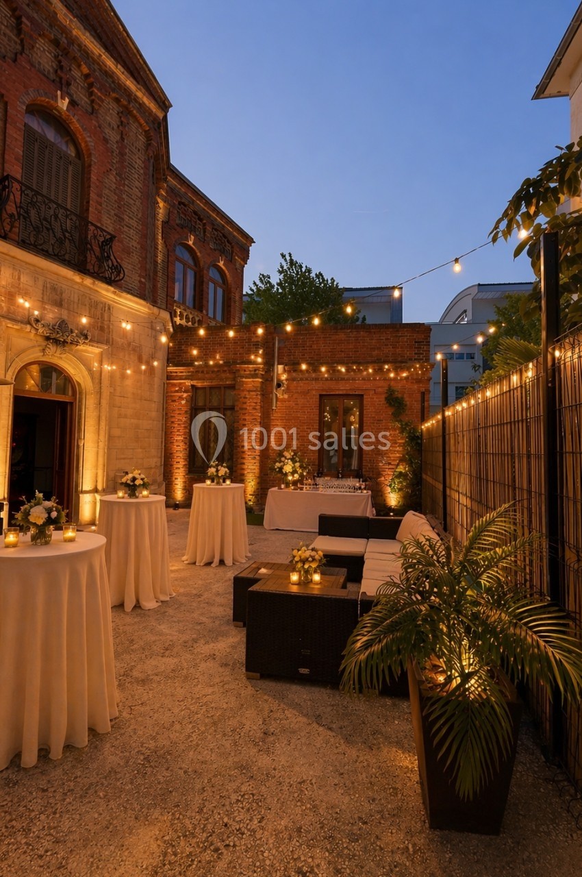 Cour extérieure éclairée par des guirlandes lumineuses, avec tables hautes décorées et mobilier de salon en soirée.