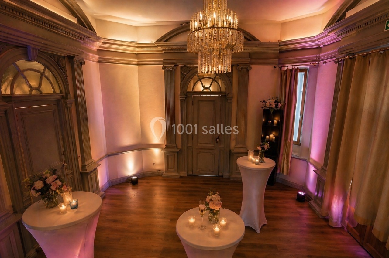 Salle élégante avec parquet, tables hautes décorées de fleurs et bougies, éclairage tamisé et lustre central.