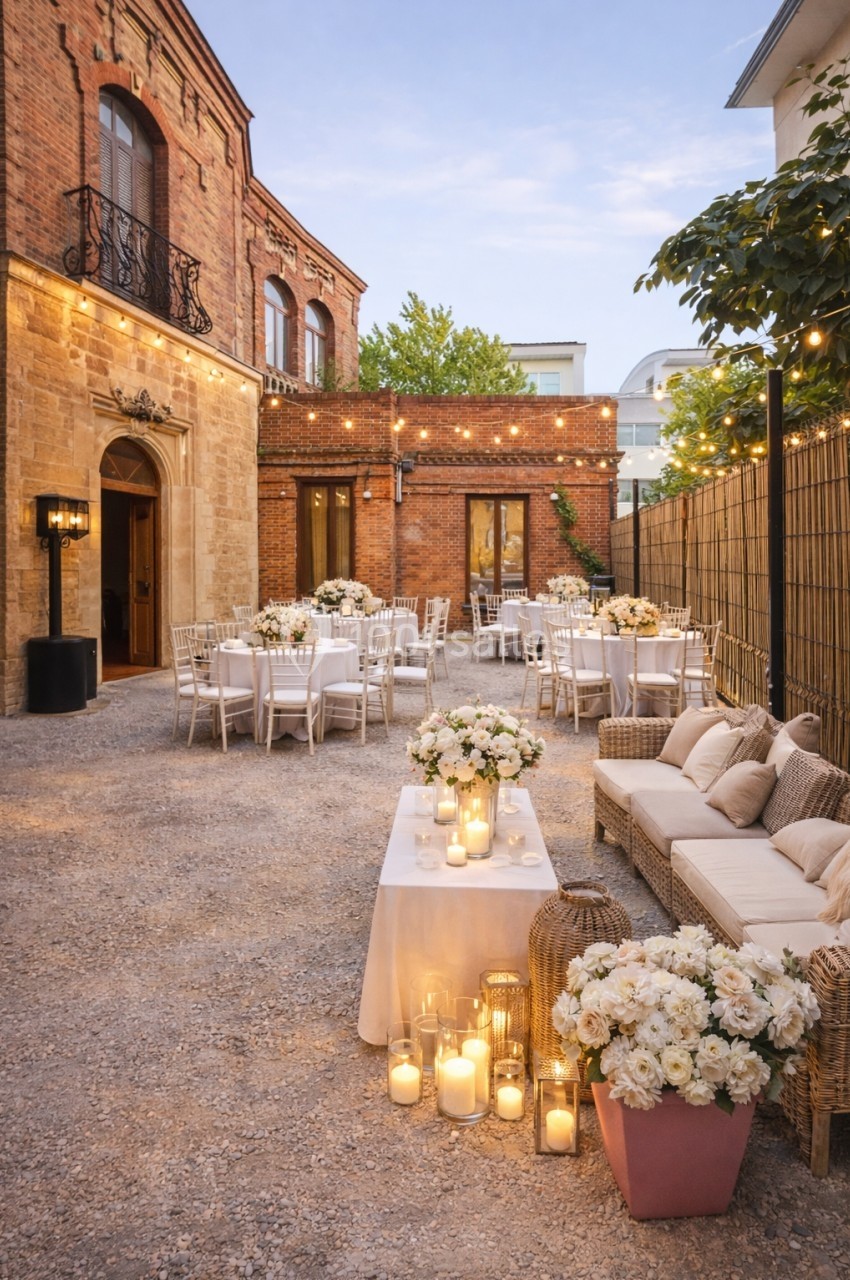 Cour extérieure aménagée pour un événement, avec tables décorées, guirlandes lumineuses et bougies entourées de fleurs…