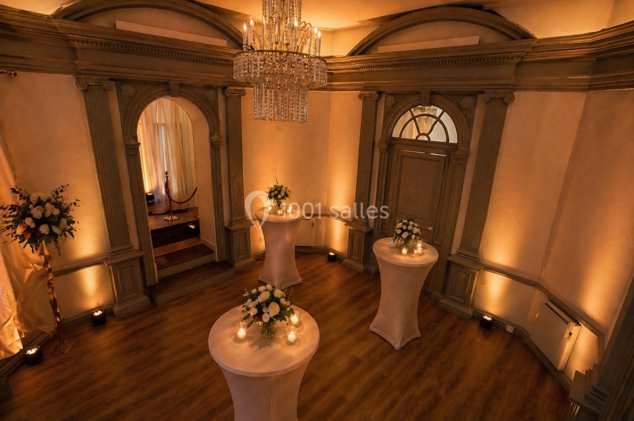 Salle élégante avec parquet, tables hautes décorées de fleurs et bougies, éclairage chaleureux et lustre central.