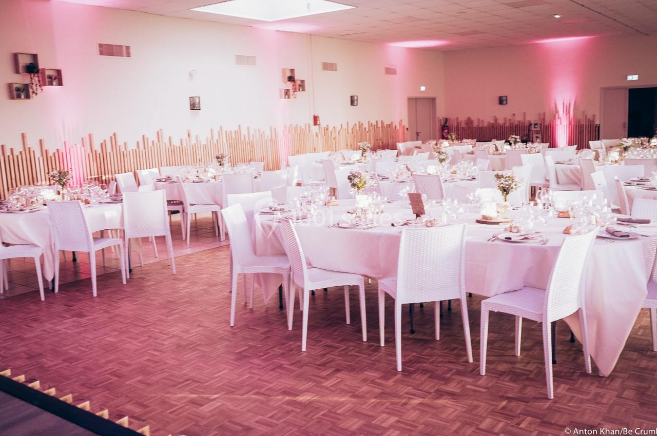 Salle de réception décorée avec des tables rondes et carrées, nappes blanches et éclairage rose tamisé.