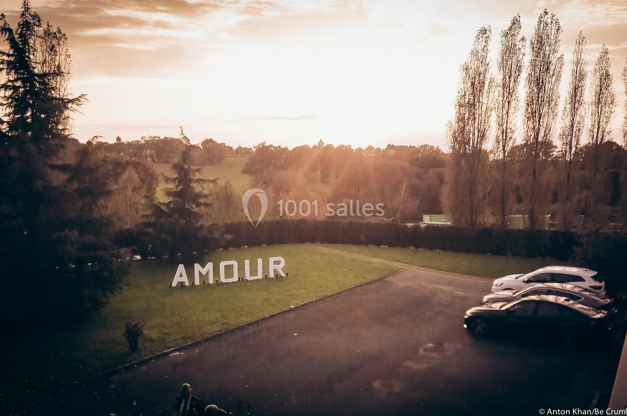Vue d'un parking bordé d'arbres au coucher du soleil, avec le mot ’AMOUR’ en grandes lettres blanches sur une pelouse.