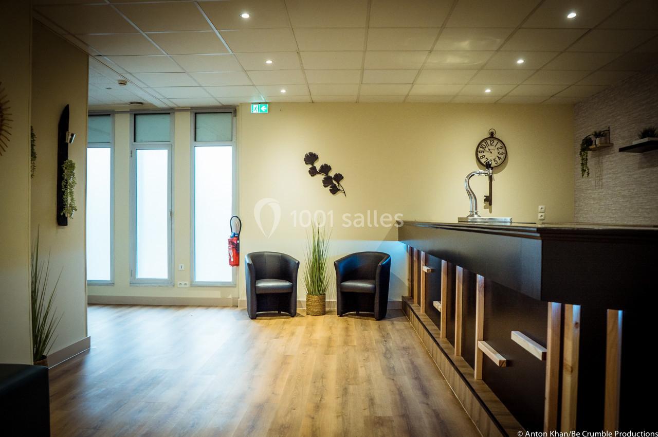 Espace d'accueil lumineux avec comptoir en bois, deux fauteuils noirs, plantes décoratives et horloge murale.