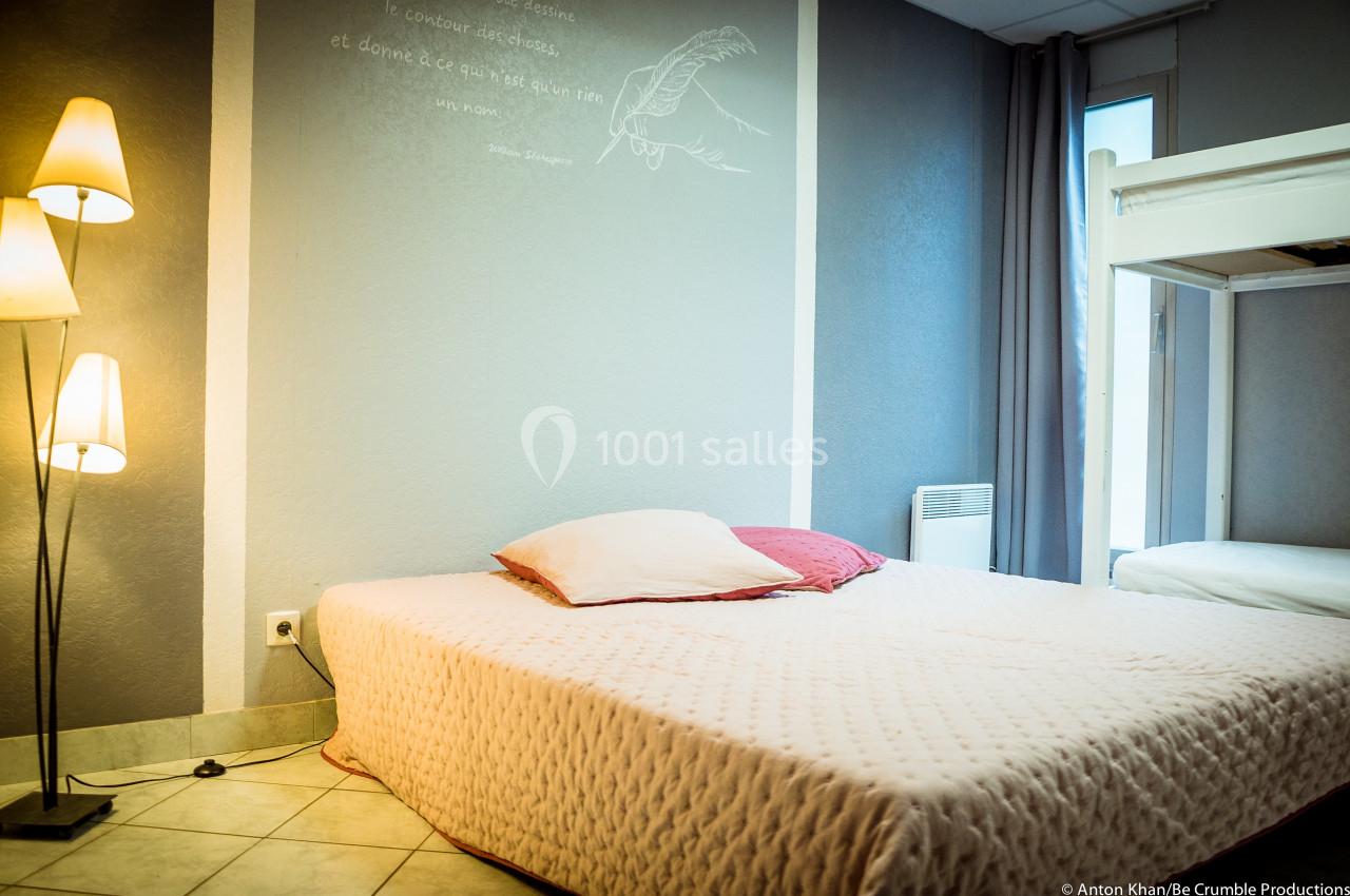 Lit double avec couvre-lit clair dans une chambre lumineuse, équipée d'une lampe sur pied et d'un lit superposé.