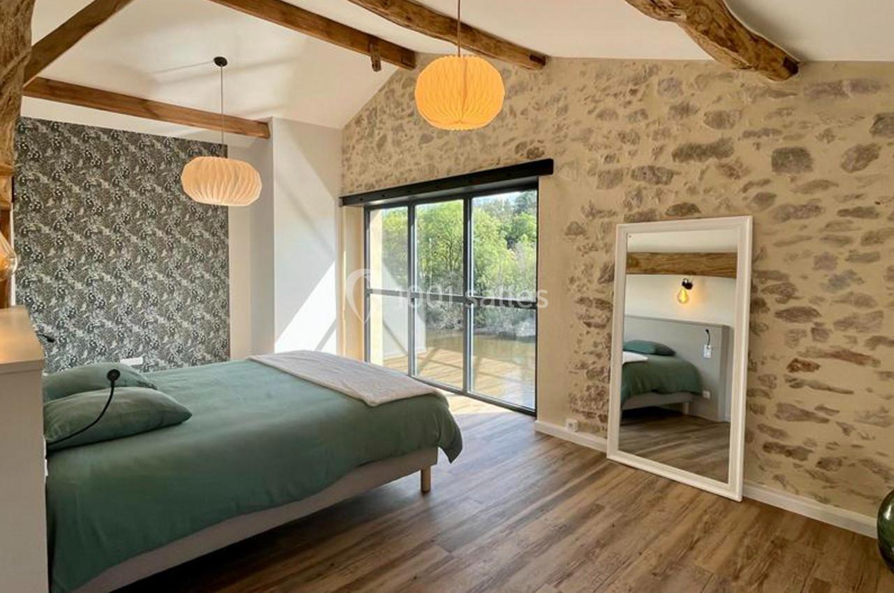 Chambre lumineuse avec lit double, mur en pierre, parquet, miroir sur pied et grande baie vitrée donnant sur l'extérieur.