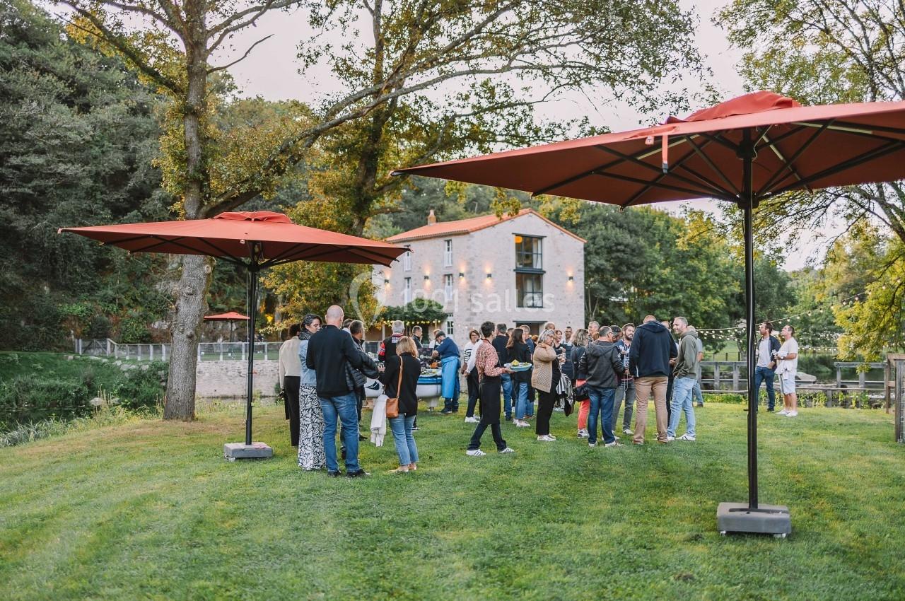 Groupe de personnes rassemblées sur une pelouse près d'un bâtiment en pierre, entouré d'arbres et de parasols rouges.