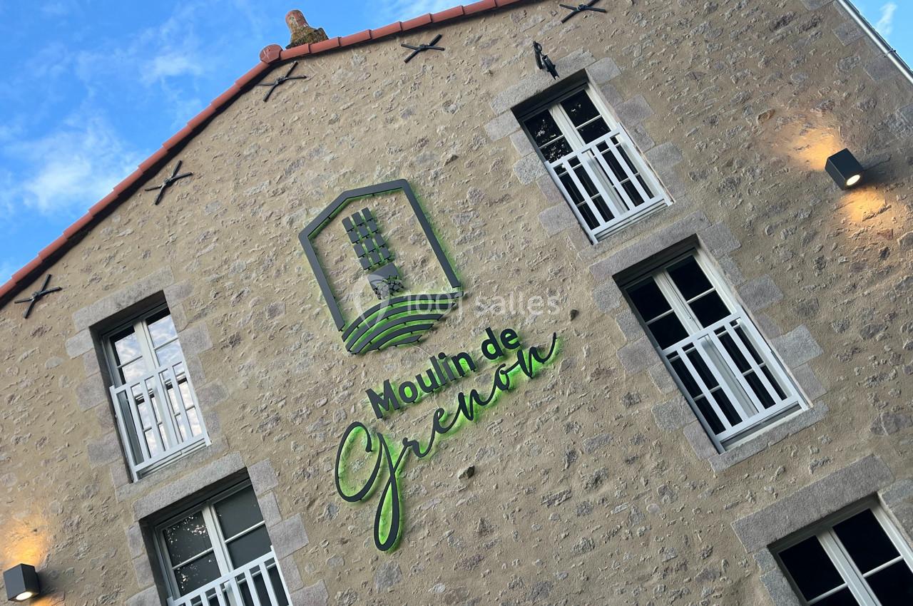 Façade en pierre d'un bâtiment avec logo ’Moulin de Grenon’ et fenêtres blanches, éclairée par des appliques murales.