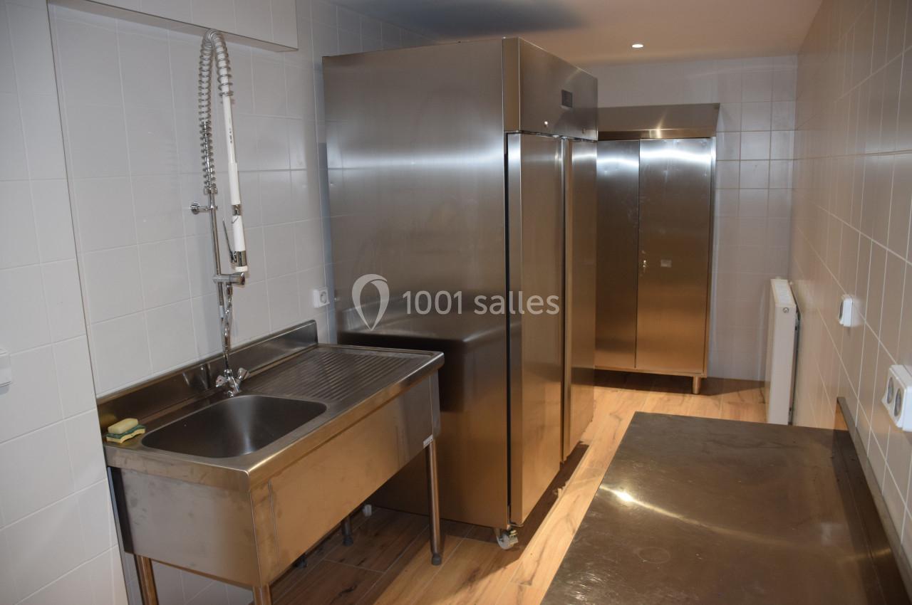 Cuisine professionnelle avec évier en inox, réfrigérateurs métalliques et murs carrelés blancs.