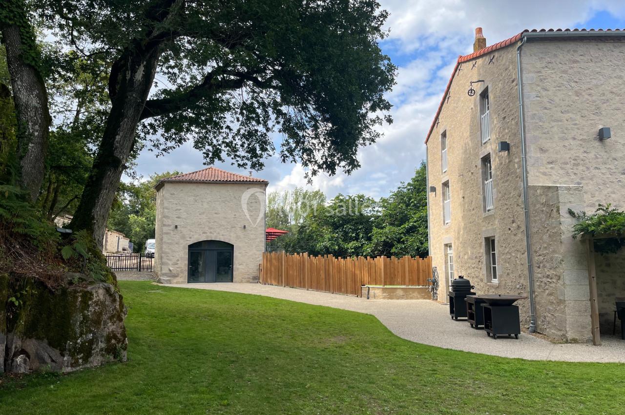 Cour avec pelouse, bâtiment en pierre, clôture en bois et arbres sous un ciel partiellement nuageux.