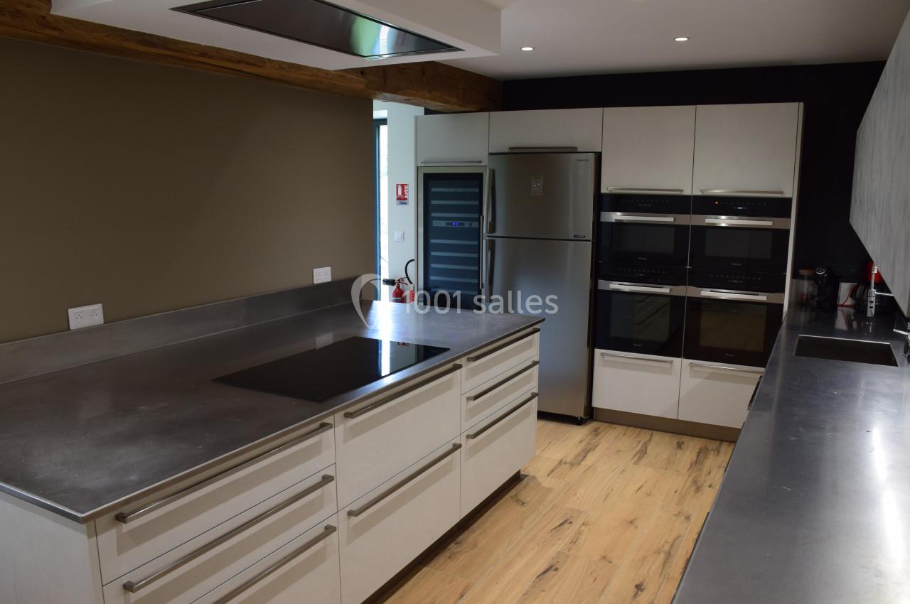 Cuisine moderne avec plan de travail en inox, plaques de cuisson, réfrigérateur, fours encastrés et sol en bois clair.