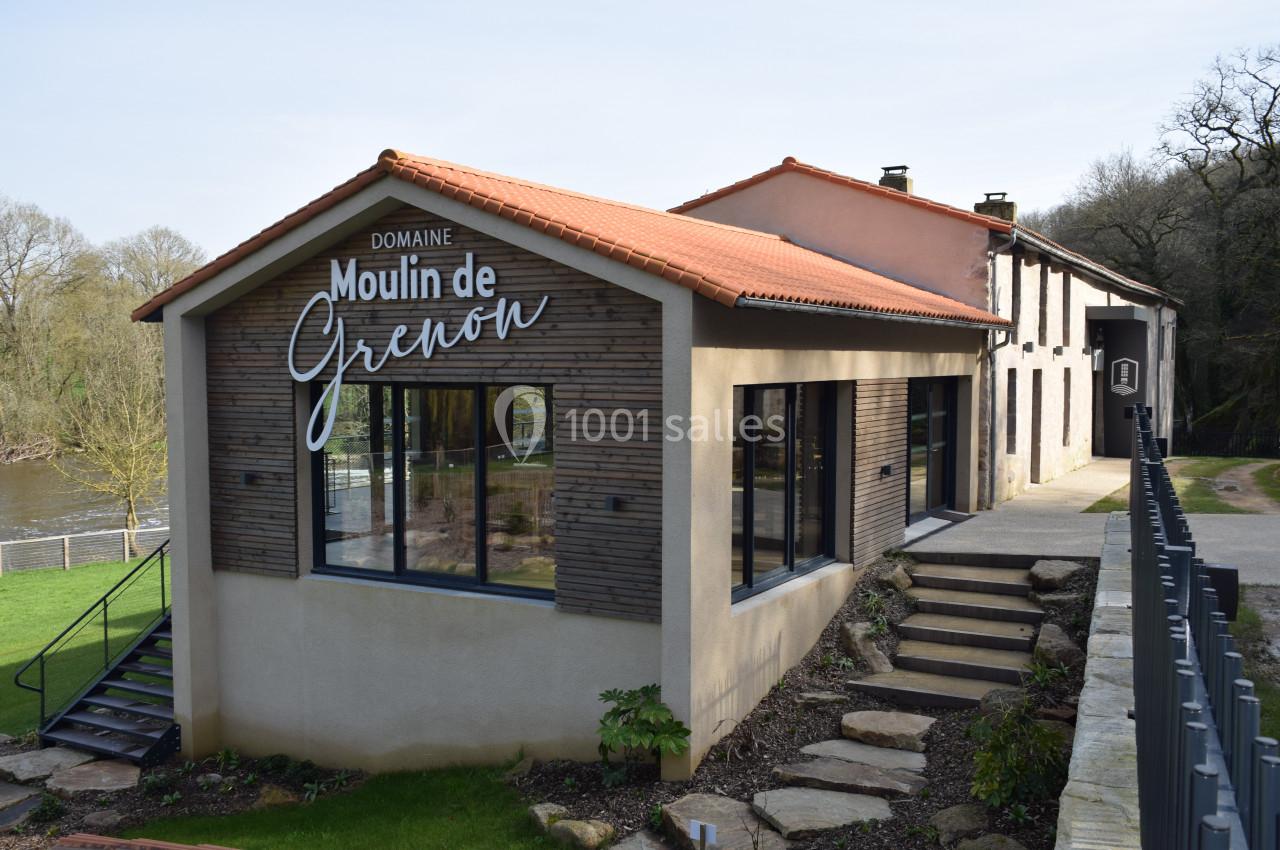 Bâtiment moderne avec façade en bois et inscription ’Moulin de Grenon’, situé près d'une rivière et entouré de verdure.