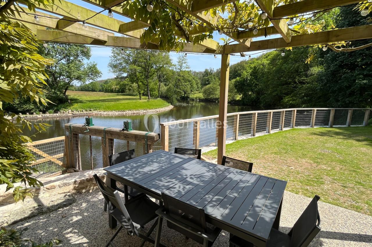 Table et chaises noires sous une pergola en bois, avec vue sur un étang entouré de verdure et une barrière en bois.