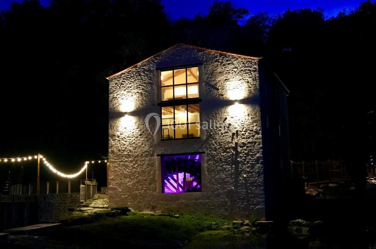 Façade d'un bâtiment en pierre éclairé la nuit, avec des lumières chaudes et une fenêtre illuminée en violet.
