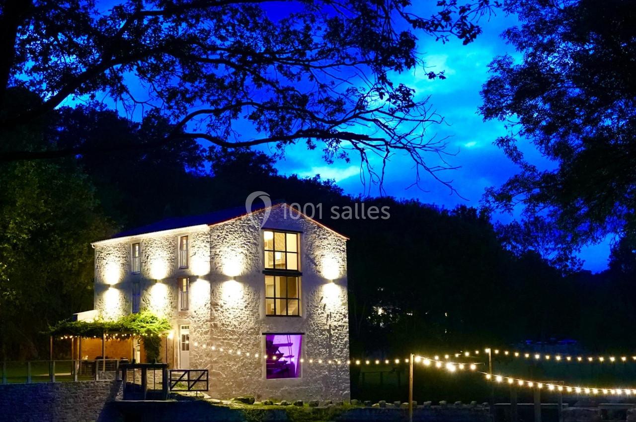 Maison en pierre éclairée la nuit, entourée d'arbres, avec des guirlandes lumineuses suspendues au premier plan.