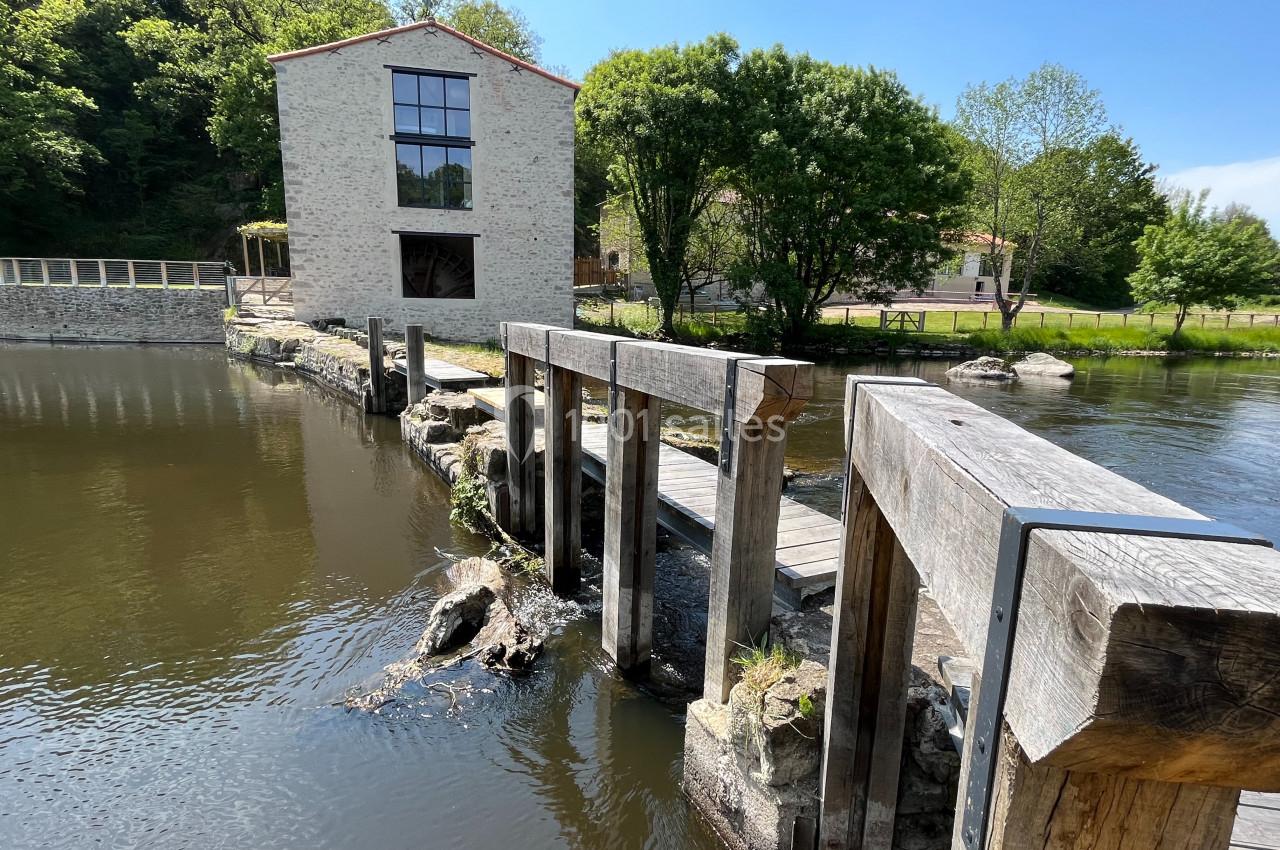 Passerelle en bois traversant une rivière avec un bâtiment en pierre et des arbres en arrière-plan.