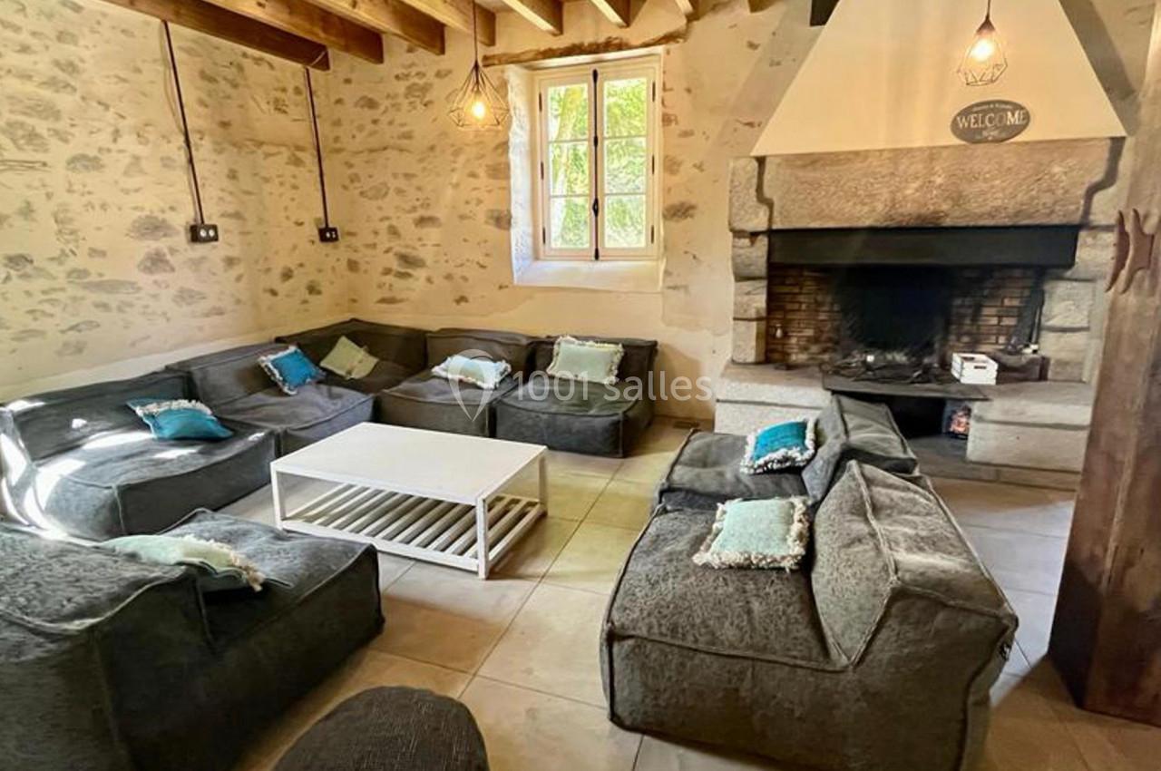 Salon rustique avec murs en pierre, poutres apparentes, cheminée en pierre et canapés gris autour d'une table basse blanche.