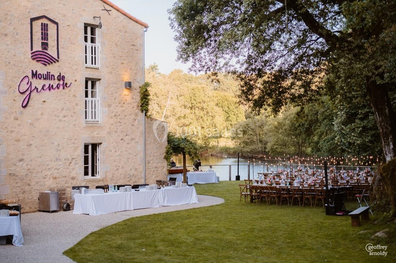 Vue d'un moulin en pierre avec une terrasse aménagée pour un événement, entouré de verdure et bordé par une rivière.