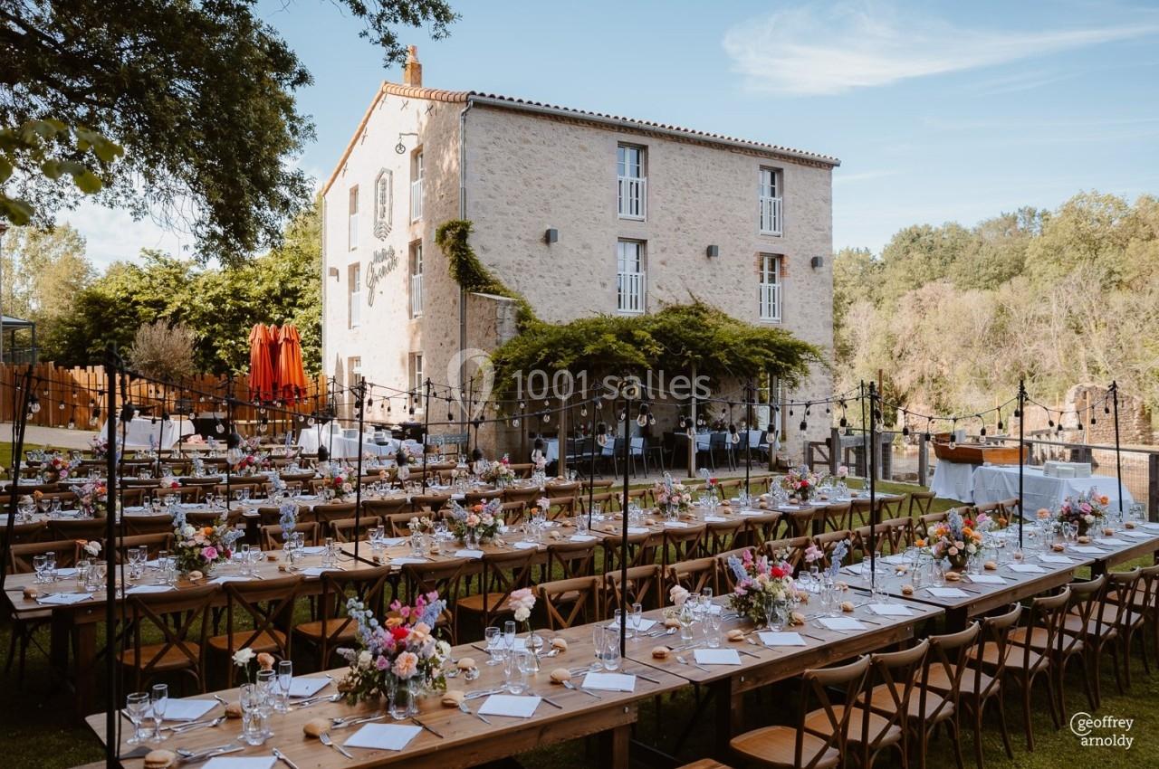 Tables en bois décorées de fleurs et dressées pour un repas en extérieur devant un bâtiment en pierre.