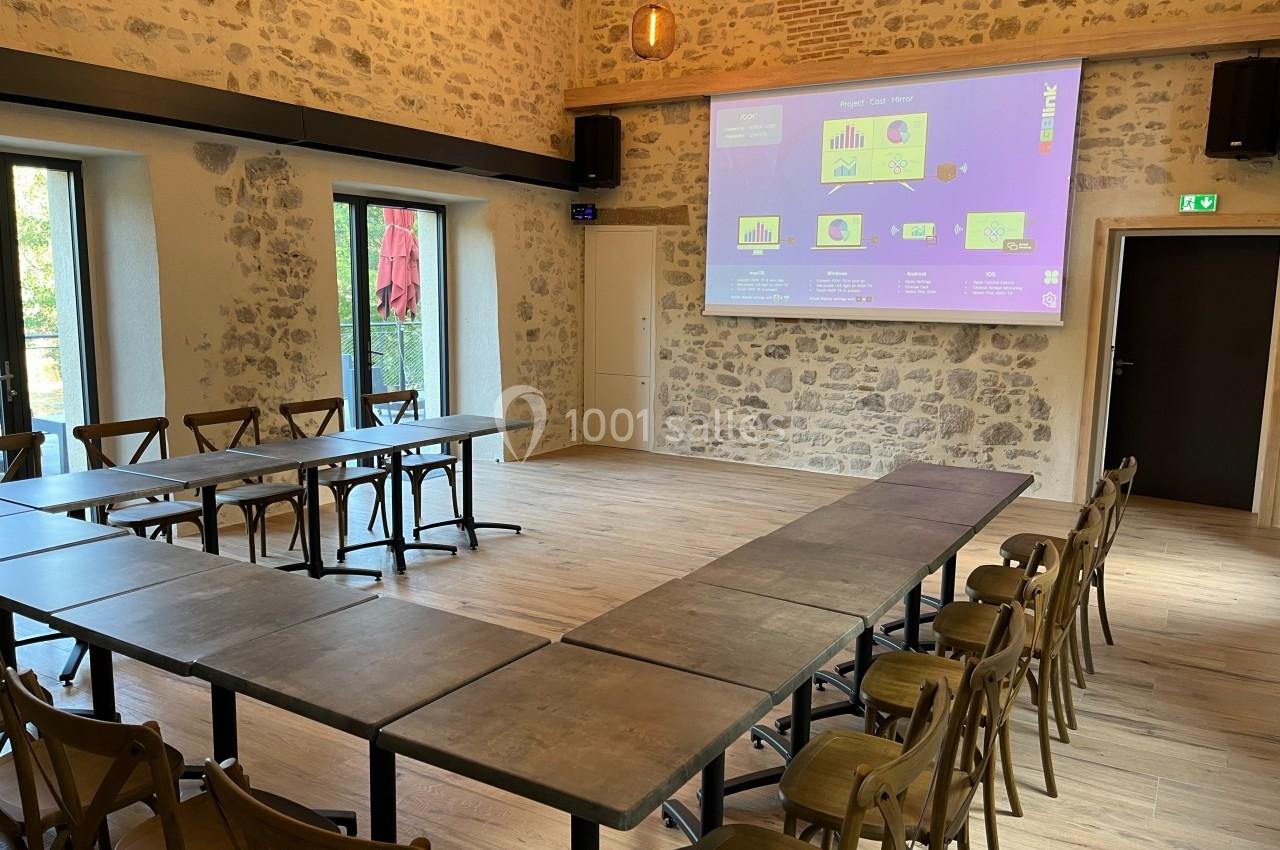 Salle de réunion lumineuse avec tables disposées en U, chaises en bois et écran de présentation projeté sur un mur en pierre.