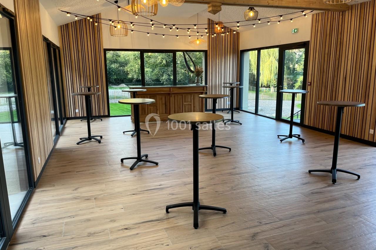 Salle lumineuse avec parquet, tables hautes noires, murs en bois et grandes baies vitrées donnant sur un jardin.