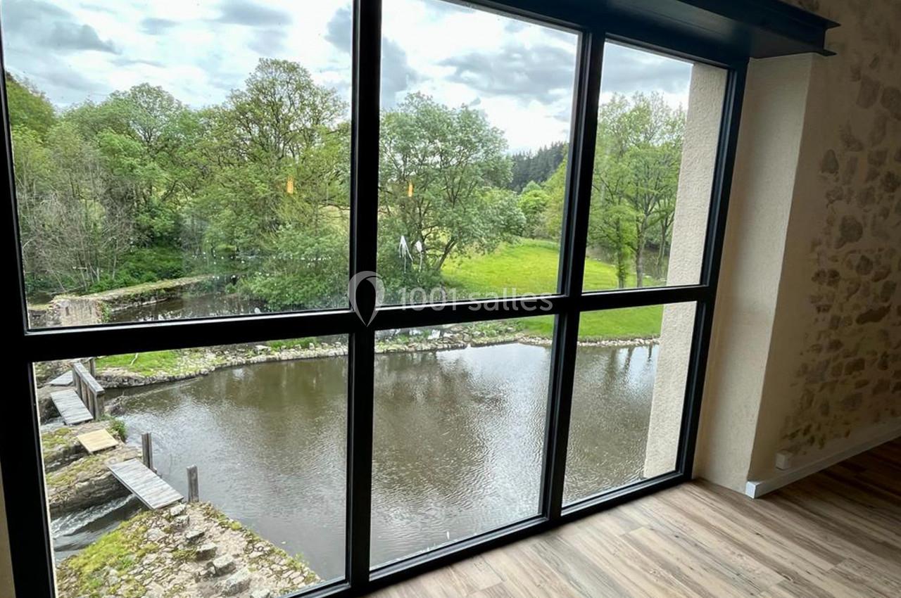 Vue sur une rivière bordée d'arbres depuis une grande baie vitrée, avec un sol en parquet à l'intérieur.