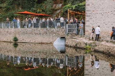 Groupe de personnes rassemblées sur une terrasse en bord de rivière, entourée de verdure et décorée de guirlandes lumineuses.