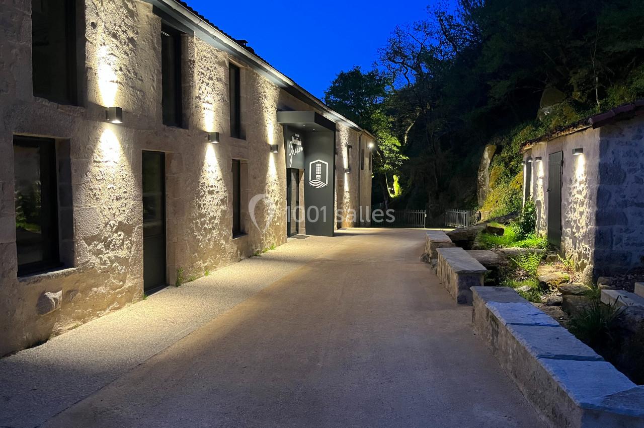 Bâtiment en pierre éclairé la nuit, avec un chemin en béton bordé de verdure et de murets.