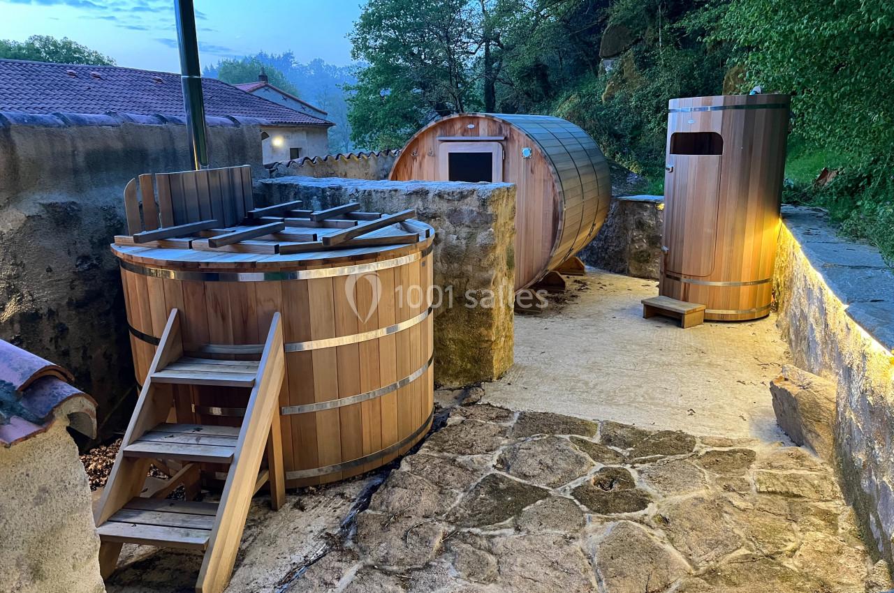 Espace extérieur avec bain nordique en bois, sauna en tonneau et douche, entouré de végétation et de pierres.