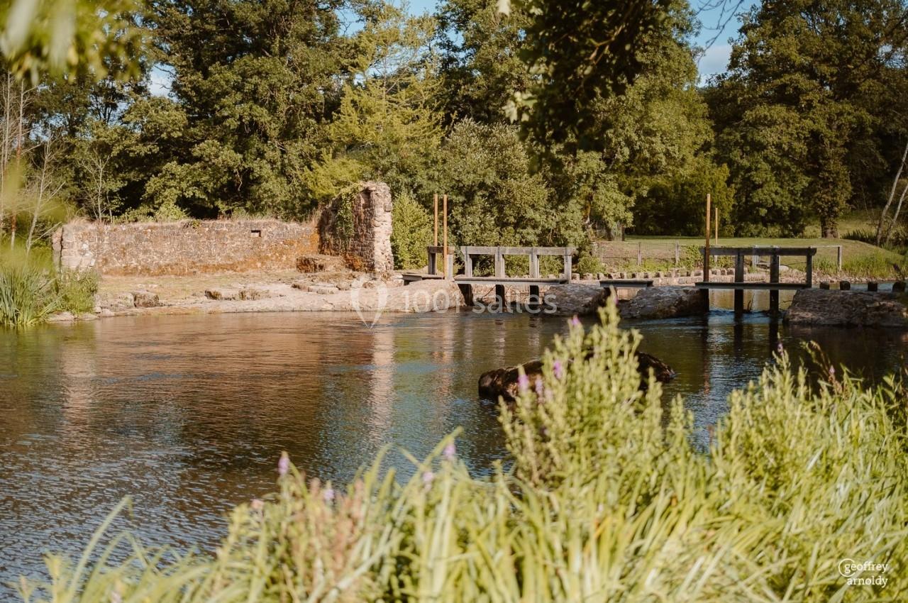 Vue d'un petit pont en bois traversant une rivière entourée de végétation et de ruines en pierre.