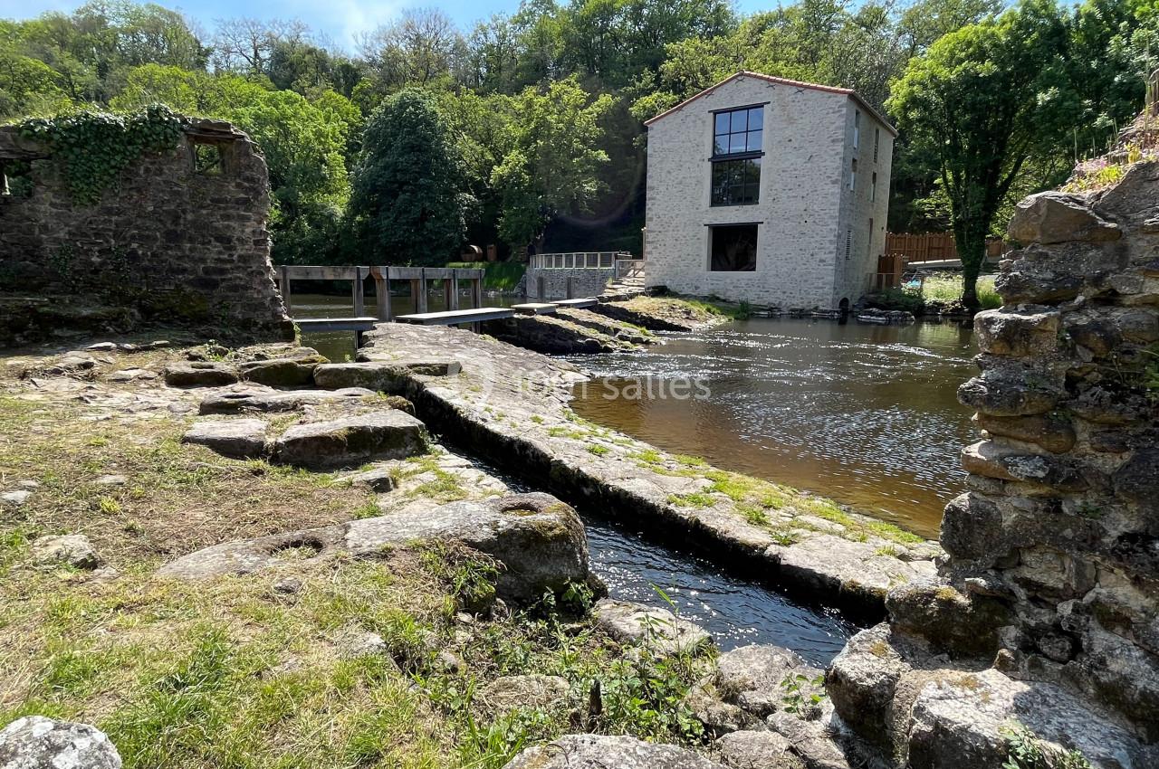 Ruines en pierre près d'une rivière avec un bâtiment rénové en arrière-plan entouré de végétation.