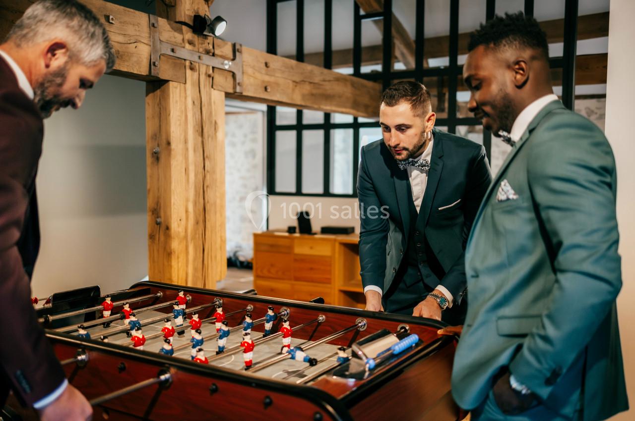 Trois hommes en costume jouent au baby-foot dans une pièce avec des poutres en bois apparentes.
