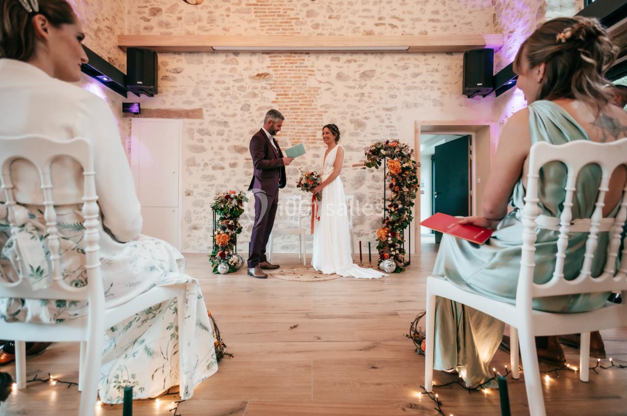 Un couple échange ses vœux devant un décor floral dans une salle en pierre, entouré de quelques invités assis.