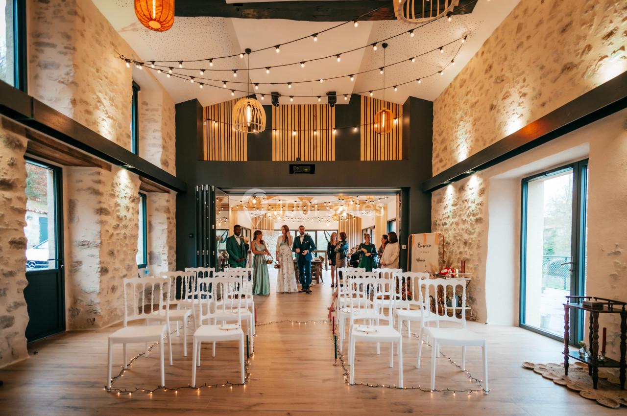 Salle de réception décorée avec des guirlandes lumineuses, des chaises blanches alignées et des invités en arrière-plan.
