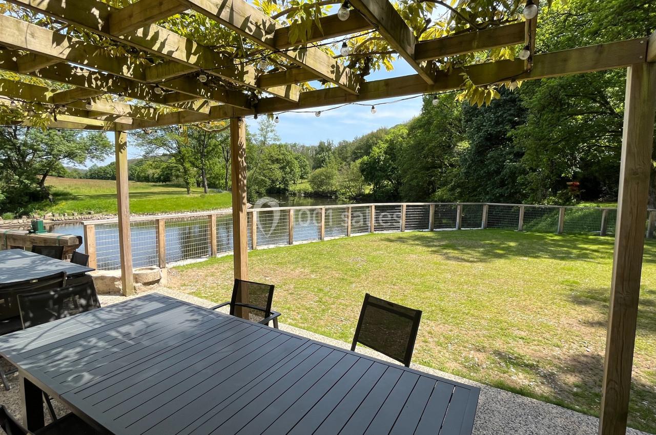 Terrasse extérieure avec pergola en bois, tables et chaises, donnant sur un jardin verdoyant et un plan d'eau.