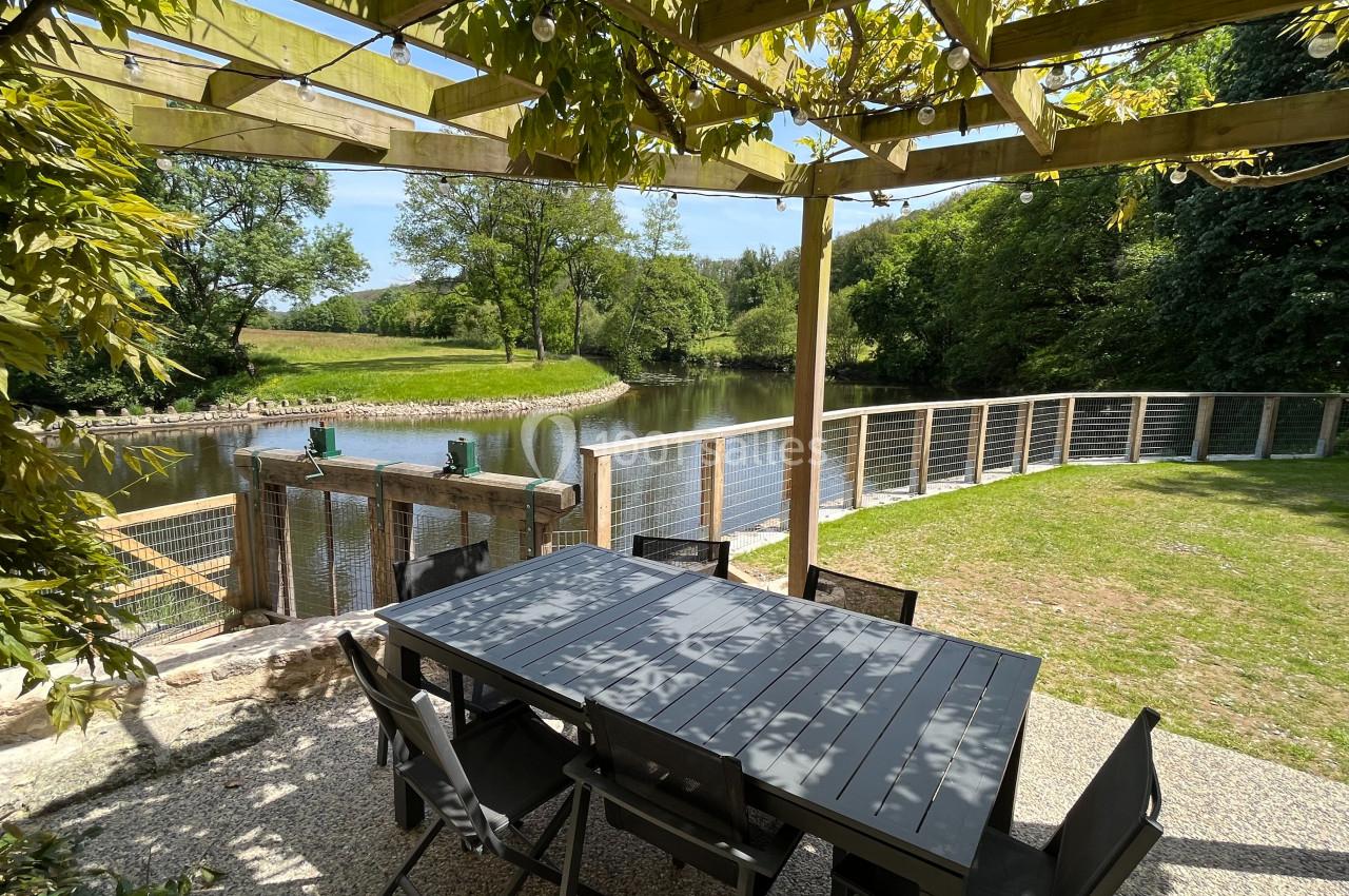 Table et chaises sous une pergola en bois, avec vue sur un étang bordé de verdure et une clôture en bois.