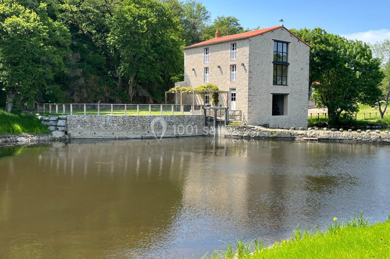 Bâtiment en pierre rénové au bord d'un étang, entouré de verdure et d'arbres sous un ciel bleu.