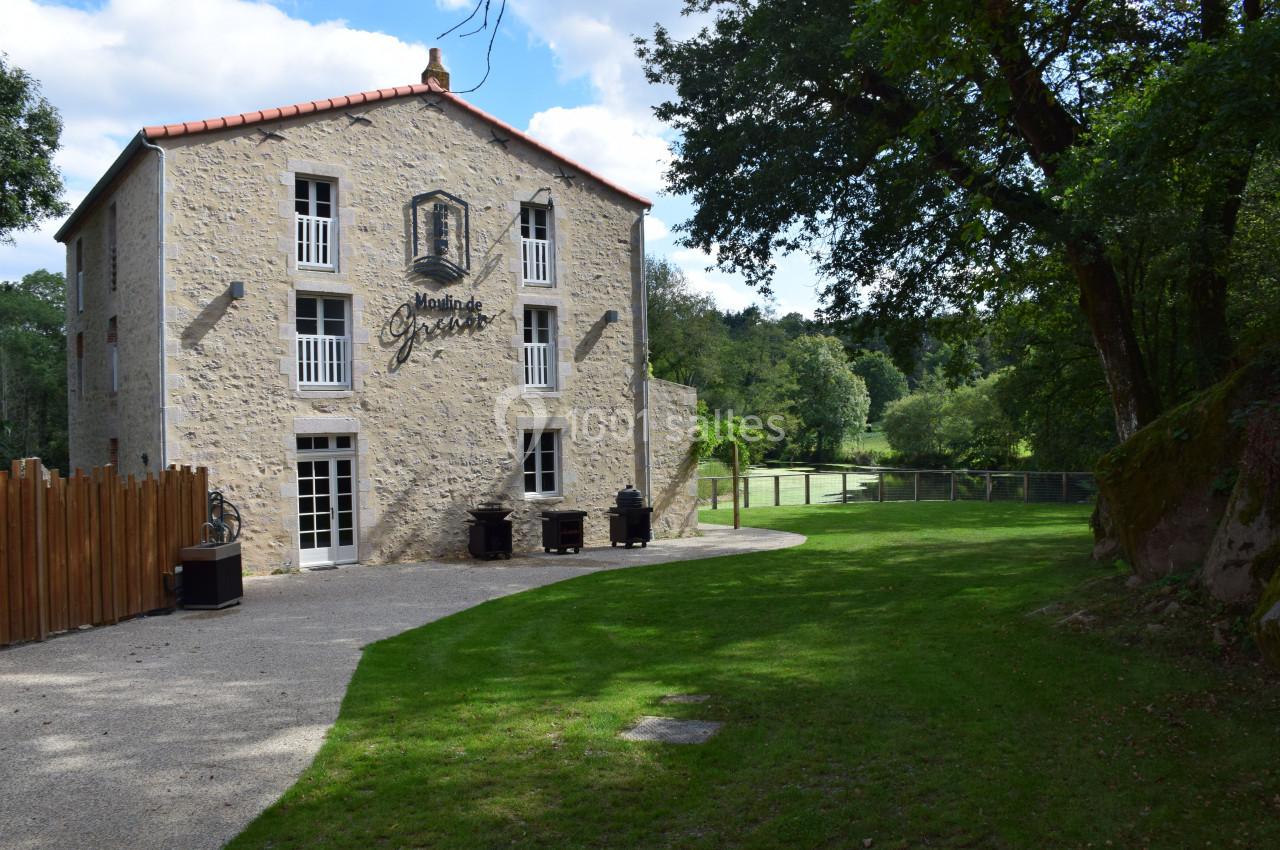 Bâtiment en pierre rénové entouré de verdure, avec une enseigne sur la façade et un chemin en gravier devant.