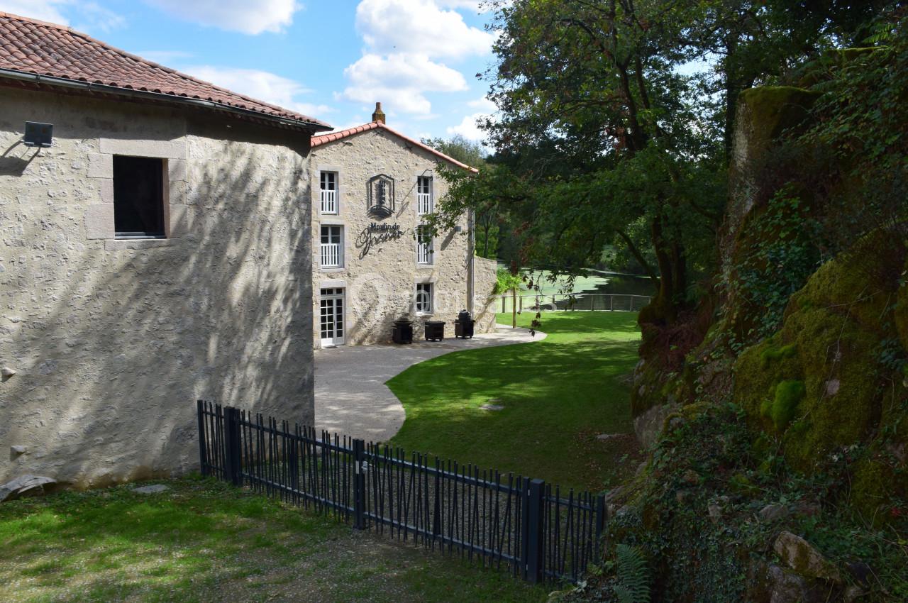 Bâtiment en pierre avec toit en tuiles, entouré d'arbres et d'une pelouse, près d'une clôture métallique.