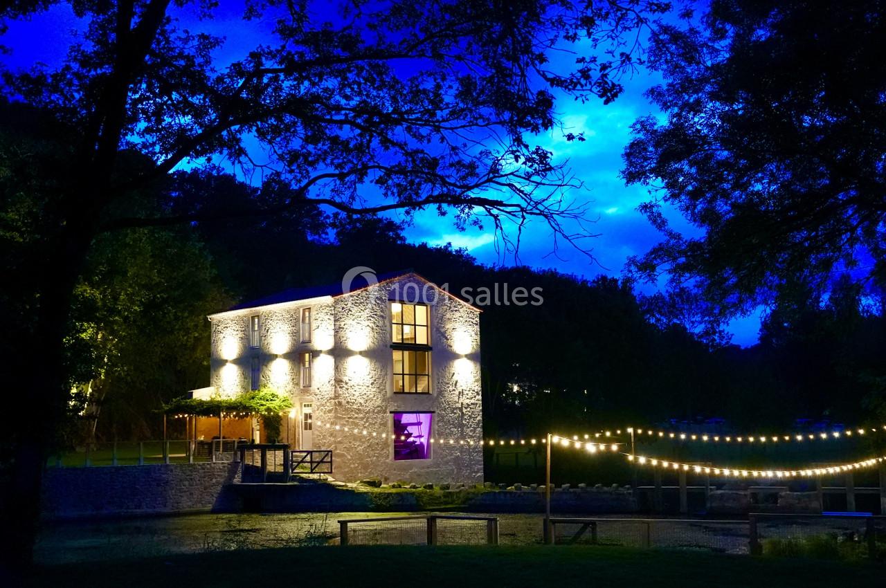 Bâtiment en pierre éclairé la nuit, entouré d'arbres et de guirlandes lumineuses près d'un cours d'eau.