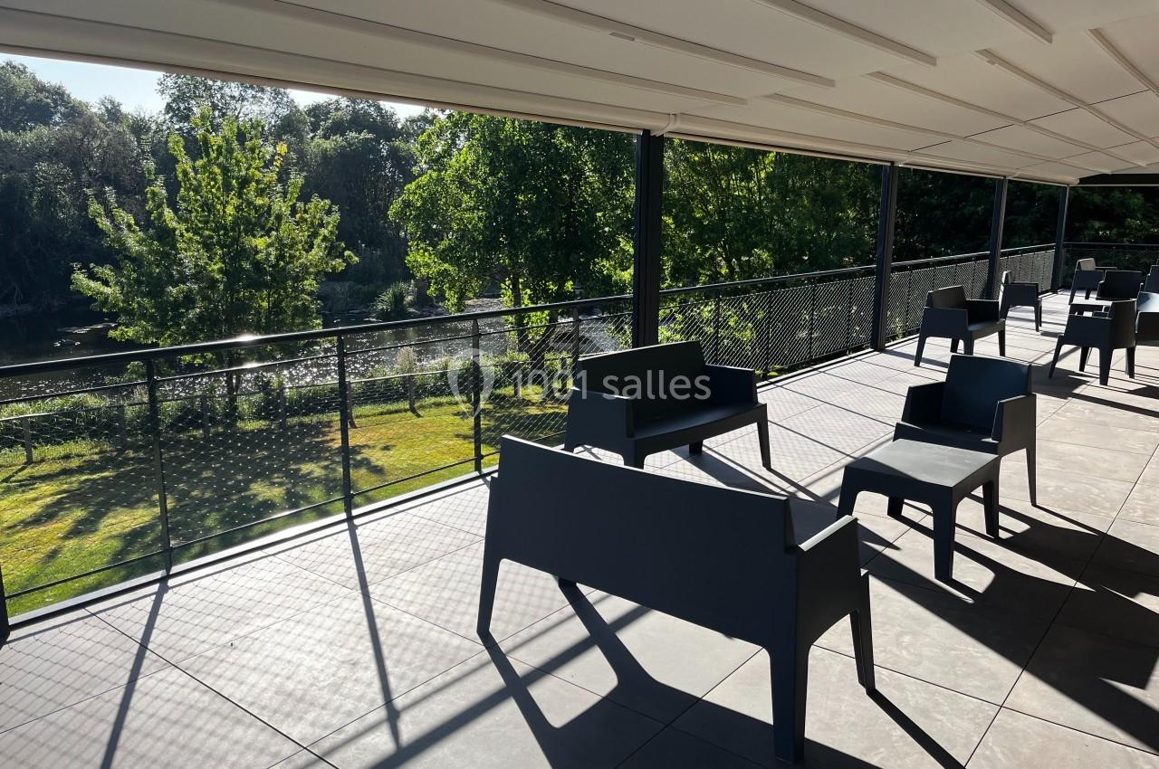 Terrasse ombragée avec des bancs noirs, vue sur un jardin verdoyant et une rivière en arrière-plan.