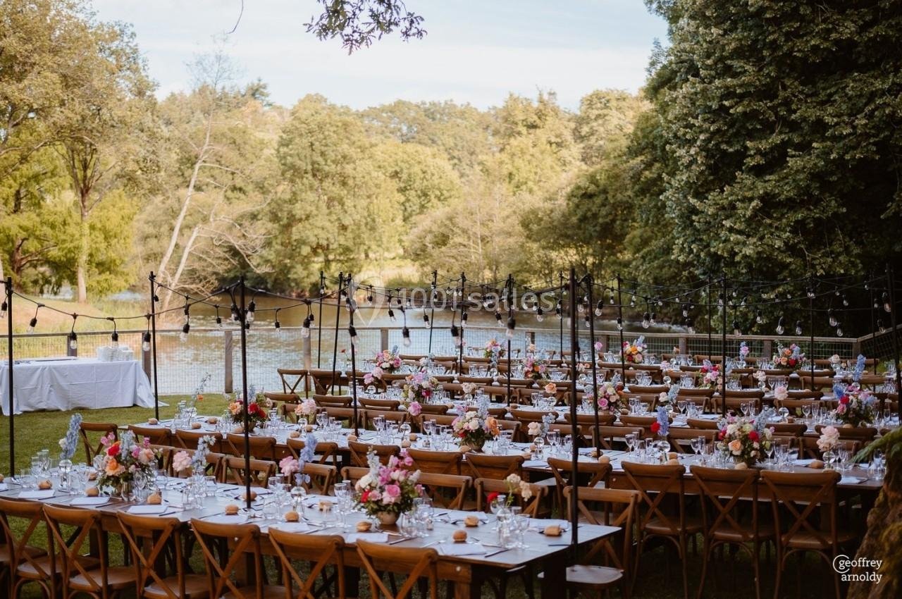 Tables en bois décorées de fleurs et vaisselle, disposées en extérieur près d'un étang entouré de verdure.